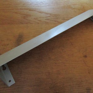 Könnte beinhalten: Ein langer, hellgrauer Regalwinkel aus Metall mit flachem, abgewinkeltem Design. Die Halterung verfügt über zwei Befestigungsenden, die jeweils mit einem runden Loch und einem schlüssellochförmigen Schlitz zur sicheren Wandmontage versehen sind.