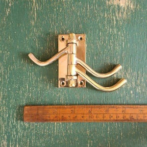 Puede incluir: Un gancho de pared de latón pulido con una placa de montaje rectangular y dos brazos giratorios. El gancho está colocado sobre una regla de madera, mostrando un ancho de aproximadamente 10 cm en la base y una envergadura total de 15 cm.