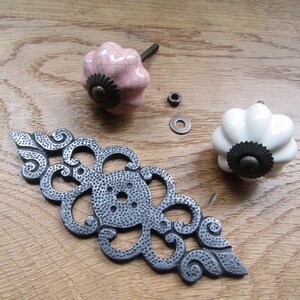K&ouml;nnte beinhalten: Zwei Keramik-Schubladenkn&ouml;pfe in geriffelter K&uuml;rbisform, einer in blassem Rosa mit Craquel&eacute;-Glasur und einer in Reinwei&szlig;. Beide haben einen dunklen Metallsockel und eine mittige Schraube. Eine dekorative, l&auml;ngliche Metall-R&uuml;ckplatte liegt bei.