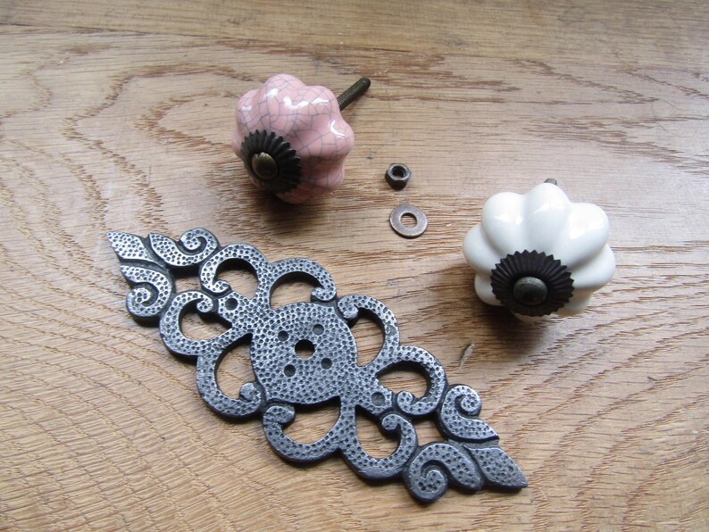 K&ouml;nnte beinhalten: Zwei Keramik-Schubladenkn&ouml;pfe in geriffelter K&uuml;rbisform, einer in blassem Rosa mit Craquel&eacute;-Glasur und einer in Reinwei&szlig;. Beide haben einen dunklen Metallsockel und eine mittige Schraube. Eine dekorative, l&auml;ngliche Metall-R&uuml;ckplatte liegt bei.