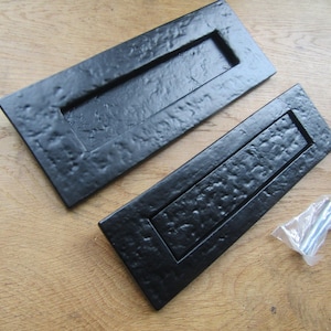 May include: Two rectangular black letter plates with a textured, hammered finish. Each plate features a central hinged flap for mail insertion. A small, clear plastic bag containing a single metal bolt and nut is positioned next to the plates.