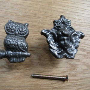 May include: Two dark grey metal cabinet knobs, one shaped like an owl perched on a branch and the other shaped like a cherub face surrounded by wings, accompanied by a single silver-toned metal mounting screw with a flat head and threaded shaft.