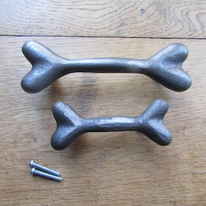 May include: Two dark grey metal drawer pulls shaped like bones, featuring a textured, hammered finish. The set includes two small silver-coloured metal screws for installation, providing a functional hardware solution for cabinets or furniture.