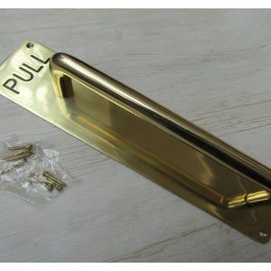 Könnte beinhalten: Eine Türzugplatte aus poliertem Messing mit rechteckiger Basis, abgerundeten Ecken und dem in schwarzen Buchstaben eingravierten Wort PULL. Das Set enthält einen langen, geschwungenen Griff sowie einen kleinen Plastikbeutel mit passenden Schrauben.