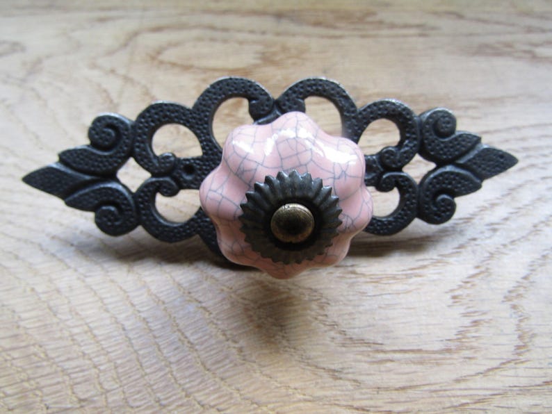 K&ouml;nnte beinhalten: Ein Schubladengriff mit einem zentralen, runden, blassrosa Keramikknopf mit dunkler Rissglasur. Der Knopf ist auf einer l&auml;nglichen, dunkelgrauen, verzierten Metallr&uuml;ckplatte mit komplexen Schn&ouml;rkeln und spitzen Enden montiert.