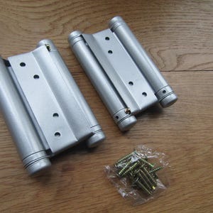 Op de afbeelding: Twee zilverkleurige dubbelwerkende veerscharnieren met cilindrische behuizingen en platte montageplaten met voorgeboorde gaten, vergezeld van een klein doorzichtig plastic zakje met een set goudkleurige metalen schroeven voor installatie.