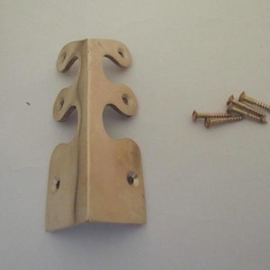 May include: A gold-toned metal corner bracket with a decorative scalloped edge and six pre-drilled screw holes. The bracket features a right-angle bend for corner reinforcement and is accompanied by a small pile of four matching gold-toned screws.