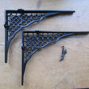 May include: Two black cast iron shelf brackets featuring an intricate lattice pattern with a circular floral motif in the corner. Each bracket has mounting holes along the vertical and horizontal edges. Four small black metal screws are included.