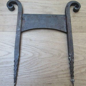 May include: A dark grey metal fireplace grate component featuring two vertical bars with curled scroll tops and pointed, textured lower ends connected by a horizontal bridge piece with a slightly curved lower edge and a weathered finish.