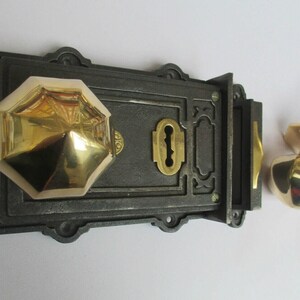 K&ouml;nnte beinhalten: Ein dunkles Gusseisen-Riegelschloss-Set mit einem polierten, achteckigen Messing-T&uuml;rknauf, der an der rechteckigen Hauptplatte befestigt ist. Ein passender, achteckiger Messing-T&uuml;rknauf mit Metallstift liegt separat auf der rechten Seite.