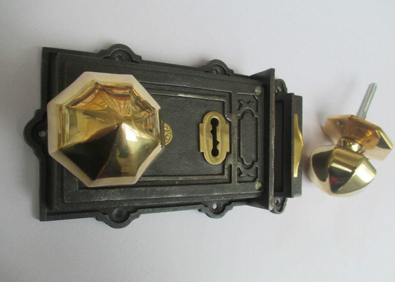 K&ouml;nnte beinhalten: Ein dunkles Gusseisen-Riegelschloss-Set mit einem polierten, achteckigen Messing-T&uuml;rknauf, der an der rechteckigen Hauptplatte befestigt ist. Ein passender, achteckiger Messing-T&uuml;rknauf mit Metallstift liegt separat auf der rechten Seite.
