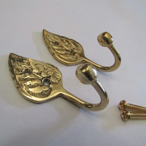 May include: Two gold-coloured metal wall hooks shaped like leaves with a curved hook end and a ball tip. Each hook features a textured leaf design with two circular mounting holes. Two small gold-coloured metal screws are included for installation.