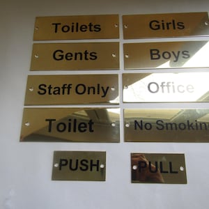 Könnte beinhalten: Eine Sammlung rechteckiger, messingfarbener Metallschilder mit schwarzer Schrift. Die Schilder tragen die Aufschriften Toilets, Girls, Gents, Boys, Staff Only, Office, Toilet, No Smoking, PUSH und PULL. Jedes Schild hat kleine Befestigungslöcher.