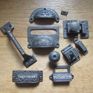 Può includere: Una collezione di ferramenta in metallo grigio scuro che include diverse maniglie per cassetti, una maniglia lunga, un pomello rotondo e un meccanismo di chiusura. Ogni pezzo presenta intricati motivi in rilievo e fori per le viti di montaggio.