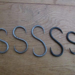 Puede incluir: Cinco ganchos de metal en forma de S dispuestos en una fila horizontal. Los ganchos varían ligeramente en tamaño y grosor, presentando un acabado metálico gris oscuro liso con un diseño curvo en ambos extremos.