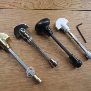 May include: Four T-shaped window handles with semi-circular grips in brass, chrome, black, and white finishes. Each handle features a long metal spindle, a small plastic washer, and a matching end cap. A small black L-shaped hex key is included.