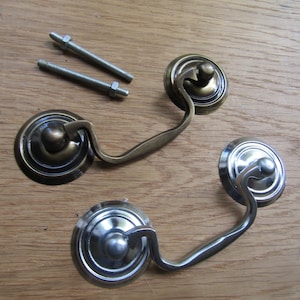 May include: Two metal drawer pulls with swinging handles and circular backplates, one in an antique brass finish and one in a polished silver finish, accompanied by two threaded metal bolts with matching nuts for installation purposes.