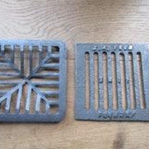 Puede incluir: Dos rejillas de drenaje cuadradas de hierro fundido gris oscuro. La rejilla izquierda presenta un patrón geométrico radiante de barras. La derecha tiene ranuras verticales paralelas y el texto en relieve J.A. FROM, S.A.I.E y FRANCE.