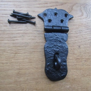 May include: A black metal gate hinge with a textured finish and a decorative spade-shaped top plate featuring five screw holes. The hinge includes a central loop and is accompanied by a small pile of five matching black metal screws for installation.