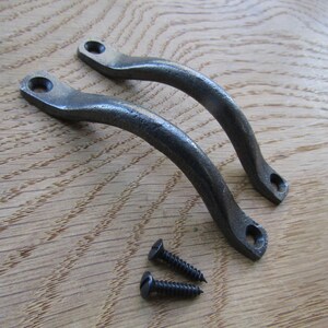 May include: Two dark grey metal drawer pulls with a curved, arched design and a textured finish. Each handle features two circular mounting holes at the ends. Two small, matching dark grey metal screws are positioned below the handles.