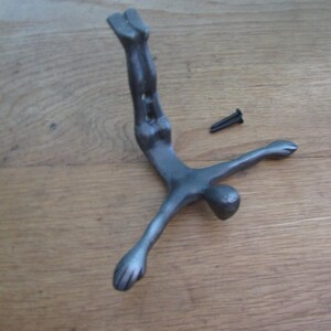 Op de afbeelding: Een donkergrijze metalen wandhaak in de vorm van een menselijk figuur in een duikpositie, met de armen gespreid en de benen omhoog gericht. De haak bevat een kleine zwarte montageschroef die naast het metalen figuur is geplaatst.
