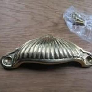 Puede incluir: Un tirador de copa de metal en tono dorado con forma semicircular, detalles acanalados verticales y dos orificios para tornillos en los extremos planos. Una pequeña bolsa de plástico transparente con dos tornillos está junto al tirador.