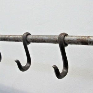 May include: A wall-mounted metal rack featuring a horizontal rod with four removable S-shaped hooks. The rack is supported by two decorative dark metal brackets on each end, each featuring a pointed finial and a star-shaped detail at the base.
