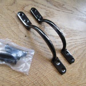 Può includere: Due maniglie ad arco in metallo nero con piastre di montaggio su ciascuna estremità, dotate di due fori per le viti preforati. Un piccolo sacchetto di plastica trasparente contenente quattro viti in metallo nero abbinate è posizionato accanto.