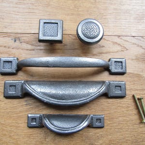 May include: A collection of dark gray metal cabinet hardware including one square knob, one round knob, one long handle, two cup pulls of different sizes, and two small metal screws. Each piece features a textured, hammered finish.