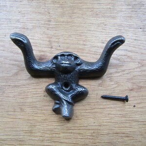 May include: A dark grey, monkey-shaped wall hook made of cast metal. The monkey is depicted with its arms extended upwards to form two hooks. A single black metal screw is positioned to the right of the monkey figure for mounting purposes.
