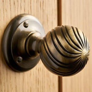 May include: A round, antique brass door knob featuring a textured, spiraling ribbed pattern that radiates from the center. The knob is attached to a circular mounting plate secured by two visible screws, showcasing a dark, metallic finish.