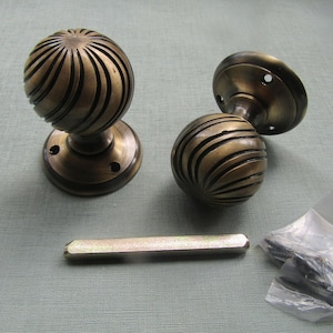 Può includere: Due pomelli per porta sferici in ottone con finitura antica scura e una texture a coste a spirale. Ogni pomello presenta una piastra di montaggio circolare con fori per le viti. Sono inclusi un perno metallico rettangolare e un sacchetto di viti.