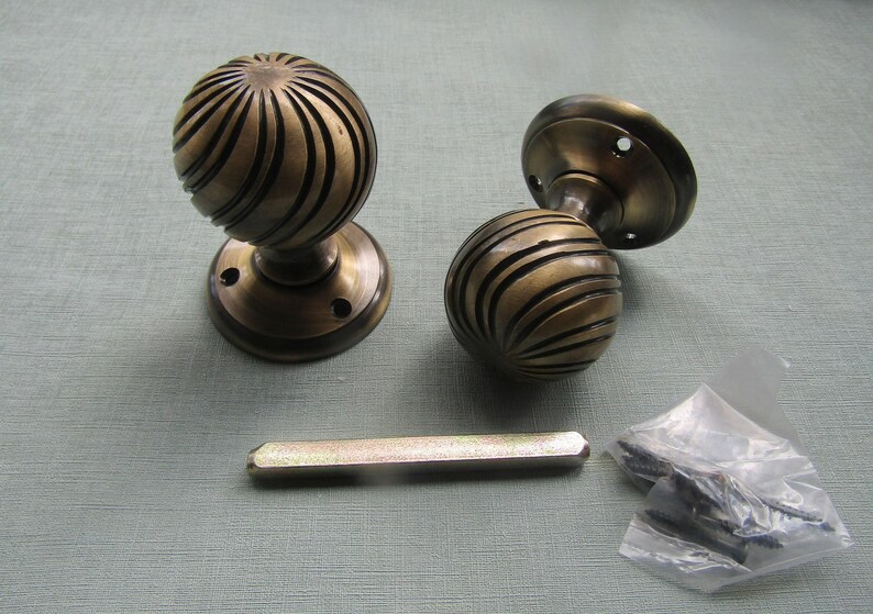 May include: Two spherical brass door knobs with a dark, antique finish and a swirling, ribbed texture. Each knob features a circular mounting plate with screw holes. A rectangular metal spindle and a small plastic bag containing screws are included.