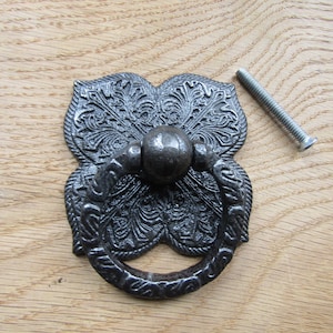 Könnte beinhalten: Ein dunkelgrauer Schubladengriff aus Metall mit einer dekorativen, verzierten Rückplatte mit komplexen, floral geprägten Mustern und einem kreisförmigen, schwingenden Ringgriff, begleitet von einer einzelnen Gewindeschraube.