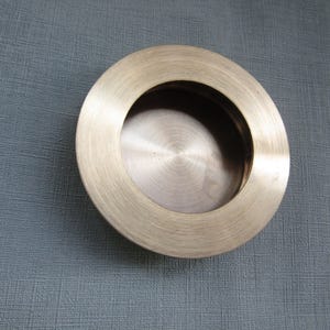 Op de afbeelding: Een ronde, geborstelde messing inbouwgreep met een gladde, metalen afwerking en een verzonken middenstuk. Het beslag heeft een brede, platte rand en een diepe, holle binnenzijde, ontworpen voor schuifdeuren of kasten met een strakke uitstraling.
