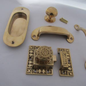 Puede incluir: Un conjunto de herrajes de latón pulido que incluye un tirador empotrado ovalado, un pomo redondo, un tirador de concha, un tirador curvo y un pestillo decorativo con base cuadrada y patrones geométricos complejos, junto con dos tornillos.