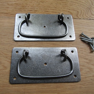 Puede incluir: Dos tiradores de cajón rectangulares de metal con acabado gris oscuro, cada uno con un asa abatible articulada unida a una placa posterior plana con cuatro orificios para tornillos en las esquinas y un orificio central, junto a tornillos plateados.