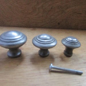 Puede incluir: Tres pomos de armario metálicos redondos de tamaños graduados, cada uno con un diseño circular concéntrico escalonado en la superficie superior. Un tornillo de montaje metálico de cabeza plana se coloca horizontalmente frente a los pomos.