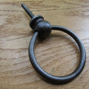 May include: A dark grey metal ring pull handle featuring a circular ring attached to a central spherical mount with a threaded screw post extending from the back for installation. The hardware has a textured, matte finish throughout its surface.