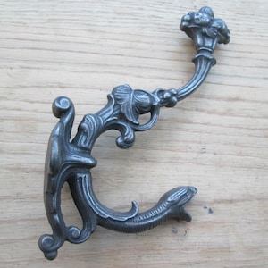Puede incluir: Un gancho de pared de metal gris oscuro con un diseño curvo y ornamentado con volutas decorativas en forma de hoja. El gancho tiene dos puntas, una que se extiende hacia arriba con un remate redondeado y texturizado.