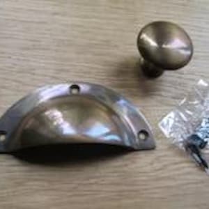 Puede incluir: Un tirador de copa de metal en tono latón con forma semicircular y dos orificios para tornillos, acompañado de un pomo de armario redondo a juego y una pequeña bolsa de plástico transparente con dos tornillos de metal para su montaje.