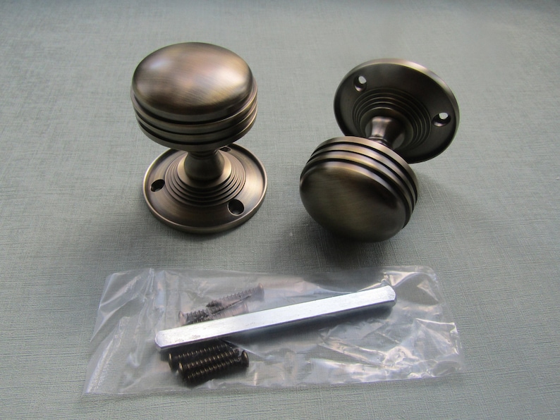 K&ouml;nnte beinhalten: Zwei runde T&uuml;rkn&ouml;pfe mit dunkelbronzenem Finish und horizontal gerippten Details am Griff. Jeder Knopf verf&uuml;gt &uuml;ber eine kreisf&ouml;rmige Montageplatte mit Schraubl&ouml;chern. Ein Plastikbeutel enth&auml;lt einen Metallstift und vier kleine Schrauben.