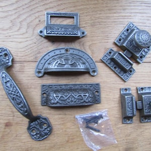 Puede incluir: Una colección de herrajes de metal gris oscuro que incluye un asa larga, un tirador de etiqueta rectangular, un tirador de media luna, un tirador rectangular, dos mecanismos de cierre y una bolsa de plástico con varios tornillos oscuros.