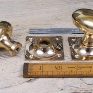May include: A set of polished brass door knobs with oval handles and square mounting plates, accompanied by a metal spindle. A wooden ruler is positioned at the bottom, showing a measurement scale in inches for the hardware components.