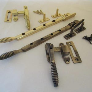 Könnte beinhalten: Ein Set aus Fensterbeschlägen, bestehend aus zwei langen Fensterfeststellern aus Metall mit gerippten Griffen, zwei passenden Fensterriegeln und kleinen Montageplatten. Das Set enthält Ausführungen in poliertem Messing und antikem Messing.