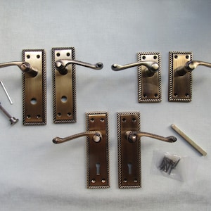 May include: A set of six antique brass door handles with rectangular backplates featuring a rope-style border. The set includes three pairs of levers, a spindle, a small metal tool, and a plastic bag containing several small mounting screws.
