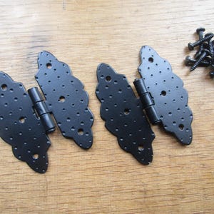 Op de afbeelding: Twee zwarte metalen scharnieren met een decoratieve geschulpte rand en een getextureerd, geput oppervlak. Elk scharnier is voorzien van meerdere voorgeboorde ronde gaten voor montage. Een kleine hoop bijpassende schroeven is inbegrepen.