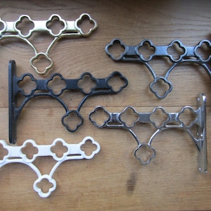 May include: A collection of five metal shelf brackets featuring a repeating quatrefoil cutout design. The set includes one polished brass, one matte black, one dark grey, one chrome, and one white bracket, each with a vertical mounting plate.