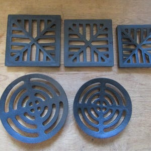 Könnte beinhalten: Eine Sammlung von fünf schwarzen Metalluntersetzern, bestehend aus drei quadratischen Teilen mit geometrischem Gittermuster und zwei kreisförmigen Teilen mit konzentrischem Ringdesign, die in zwei Reihen angeordnet sind.