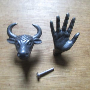 May include: Two dark grey metal cabinet knobs, one shaped like a bull head with curved horns and the other shaped like an open human hand with fingers extended. A single silver-coloured metal mounting screw is positioned below the two knobs.